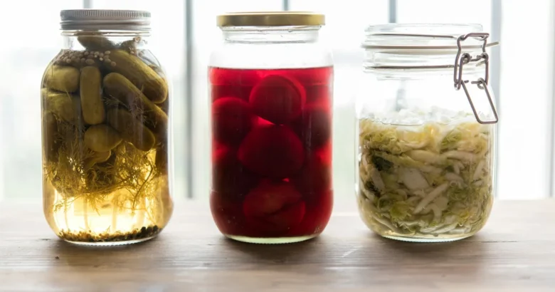 Três potes de vidro com conservas fermentadas de pepino, beterraba e repolho sobre uma mesa de madeira