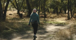 Mulher de gorro rosa, blusa azul-esverdeada e legging preta caminhando de costas numa trilha de terra entre árvores
