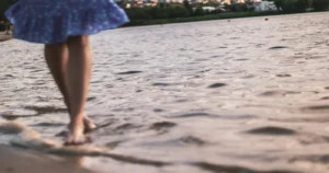 Pernas de mulher de vestido azul caminhando descalça na beira da água, com a cidade ao fundo